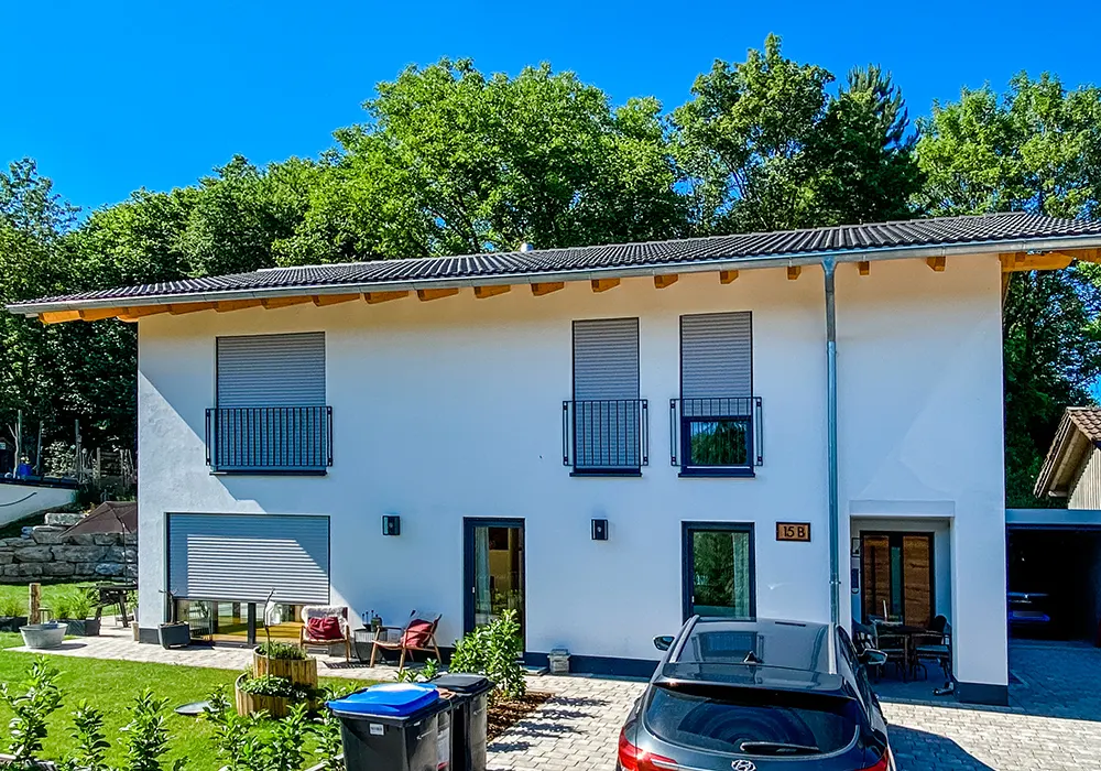 Weißes Haus mit Rollläden, Balkongittern, Sitzbereich und Auto vor der Tür, grüne Bäume im Hintergrund.