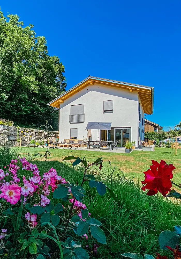 Haus mit Terrasse im Garten, im Vordergrund blühende Rosen und Rasenfläche vor blauem Himmel.