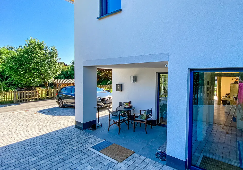 Überdachter Eingangsbereich mit Sitzgruppe und Tür, davor gepflasterter Hof mit parkendem Auto und Gartenzaun.