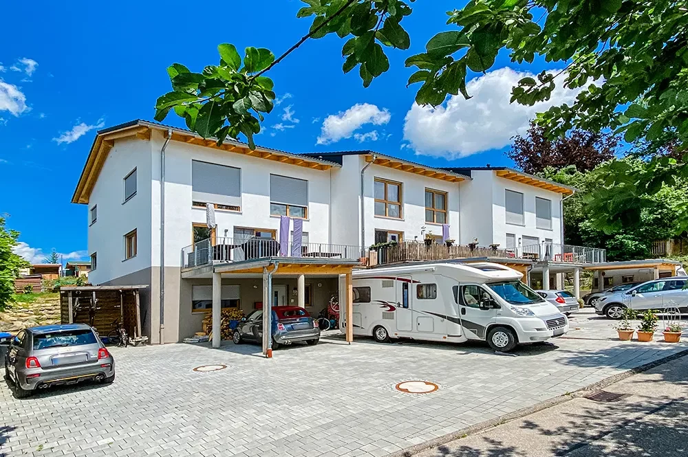 Reihenhäuser mit Balkonen und Carports, davor mehrere Autos und ein Wohnmobil auf gepflastertem Hof.