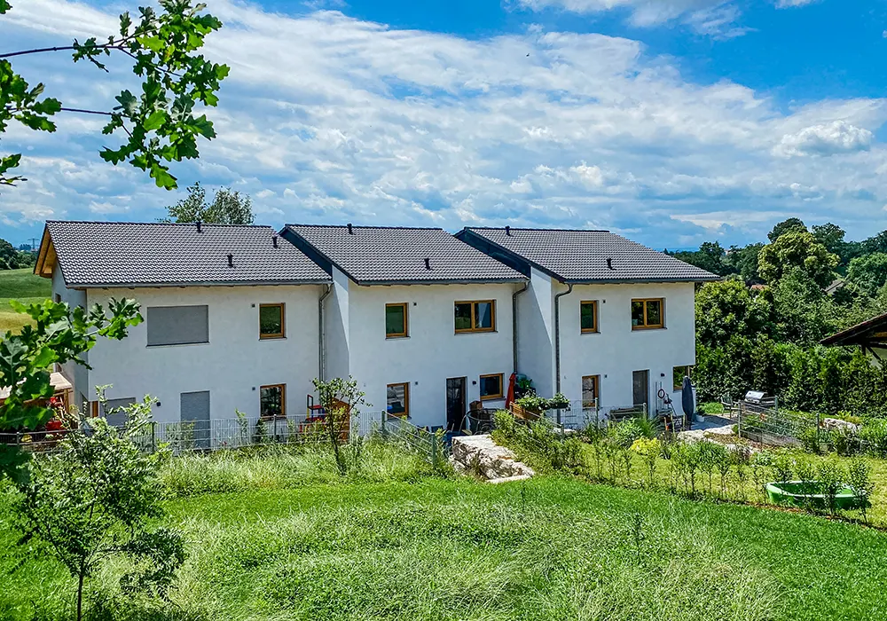 Drei verbundene Reihenhäuser mit Gärten, dahinter Felder und Bäume unter teils bewölktem Himmel.