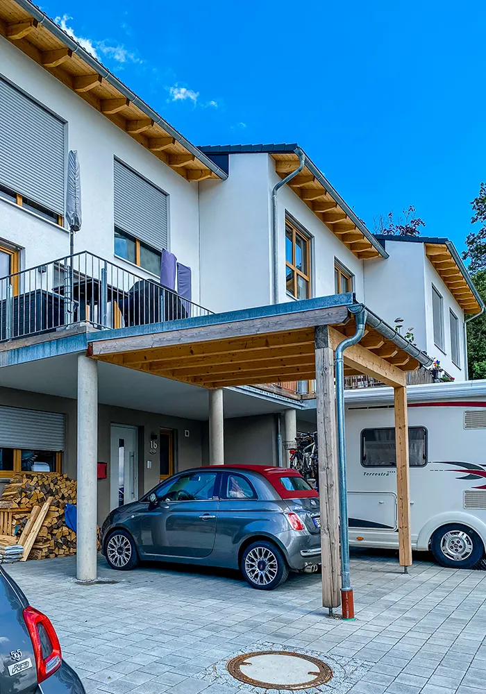Haus mit Balkon, Carport, geparktem Kleinwagen und Wohnmobil, Holzstapel neben dem Eingang.