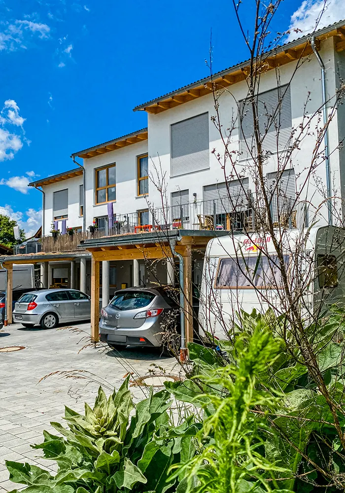 Reihenhäuser mit Balkonen, darunter Carports mit Autos und Wohnwagen, im Vordergrund grüne Pflanzen.