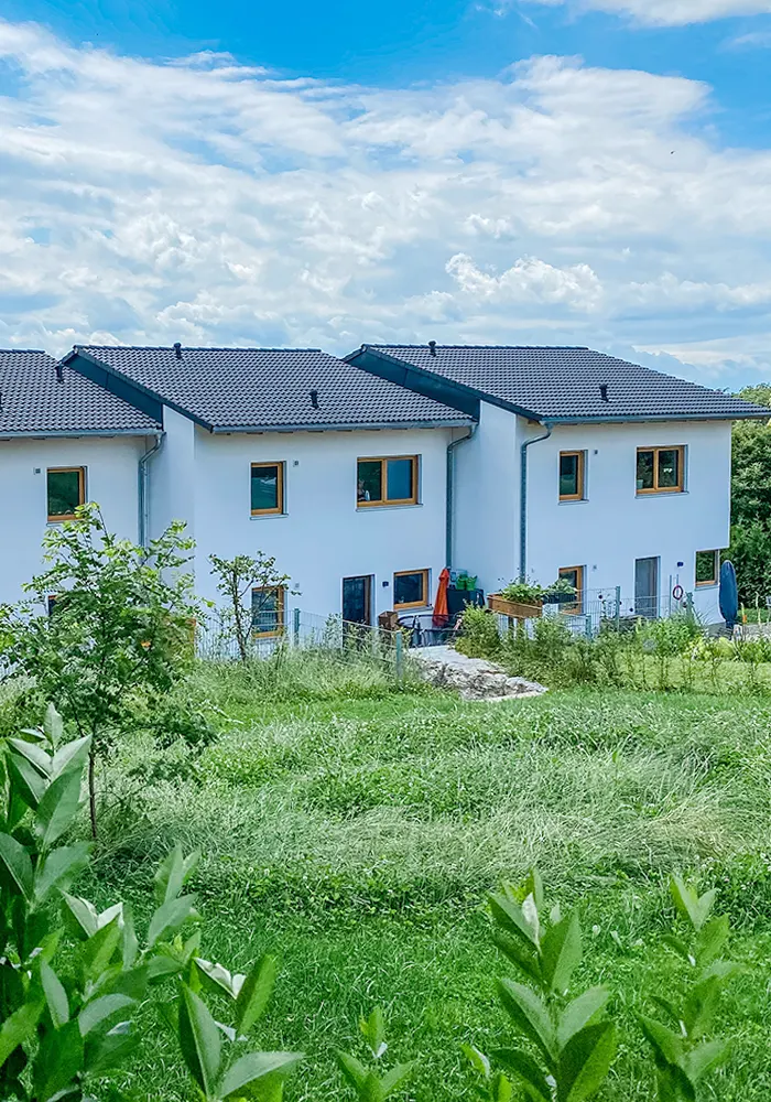 Drei moderne Reihenhäuser mit Gärten, umgeben von Wiese und Bäumen unter leicht bewölktem Himmel.