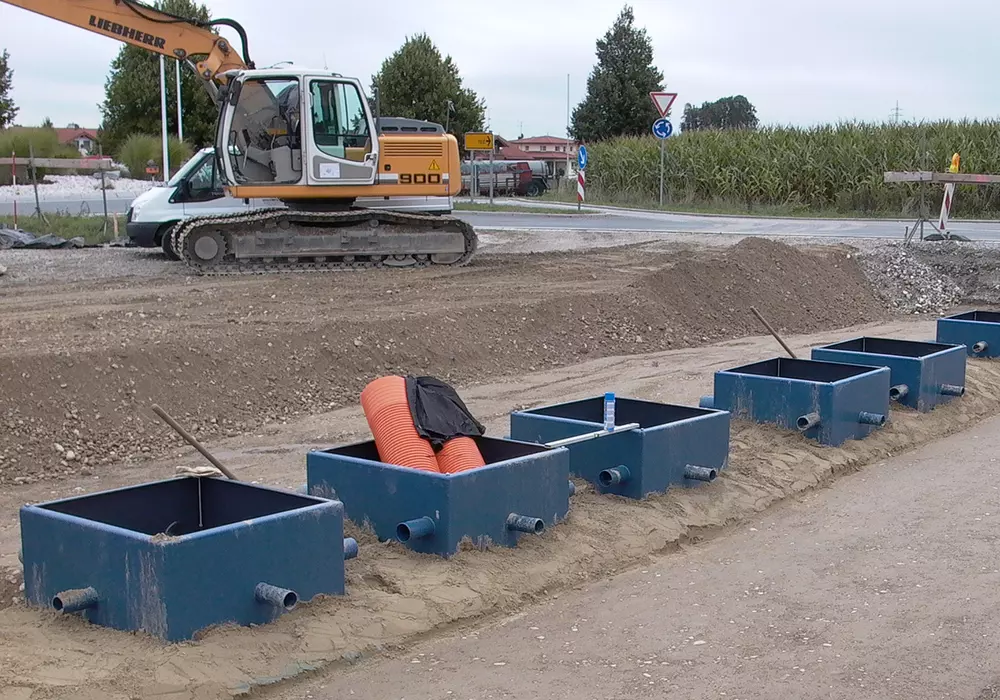 Reihe offener blauer Tankzugänge mit Rohren auf Baustelle, daneben Bagger und Verkehrsschilder im Hintergrund.