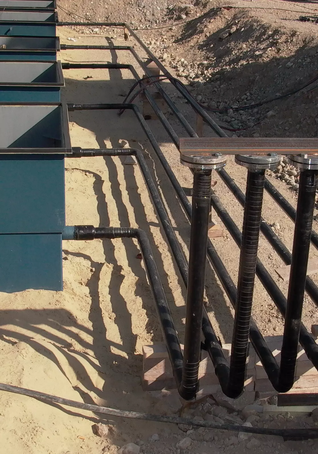 Schwarze Rohrleitungen auf Baustelle, verbunden mit blauen Tanköffnungen, verlaufen geordnet über sandigen Untergrund.