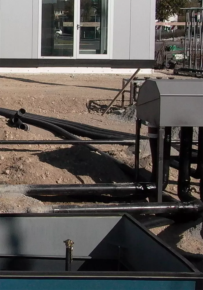 Schwarze Rohrleitungen verlaufen über Erdreich zwischen Technikbox und Gebäudefassade mit Tür und Fenster.