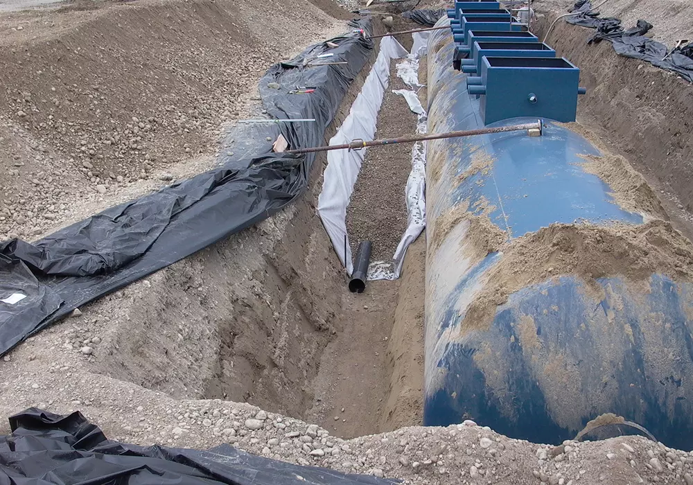 Großer blauer Erdtank in Baugrube mit Kiesbett, Rohrleitungen und schwarzer Folie zur Abdichtung.
