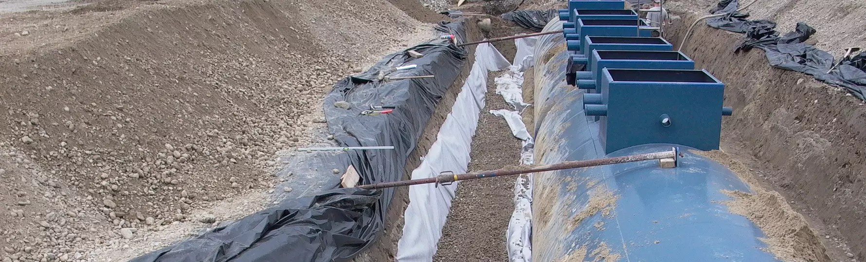 Großer blauer Erdtank in Baugrube mit Kiesbett, Rohrleitungen und schwarzer Folie zur Abdichtung.