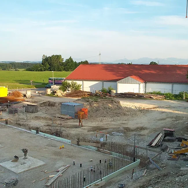 Baustelle mit Kran, Baumaterialien und Maschinen, im Hintergrund Halle mit rotem Dach und grüne Landschaft.
