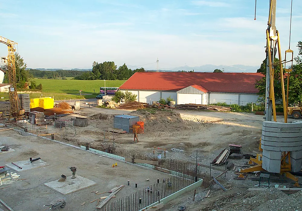 Baustelle mit Kran, Baumaterialien und Maschinen, im Hintergrund Halle mit rotem Dach und grüne Landschaft.