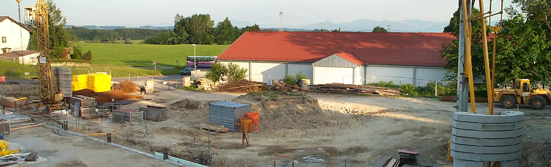 Baustelle mit Kran, Baumaterialien und Maschinen, im Hintergrund Halle mit rotem Dach und grüne Landschaft.