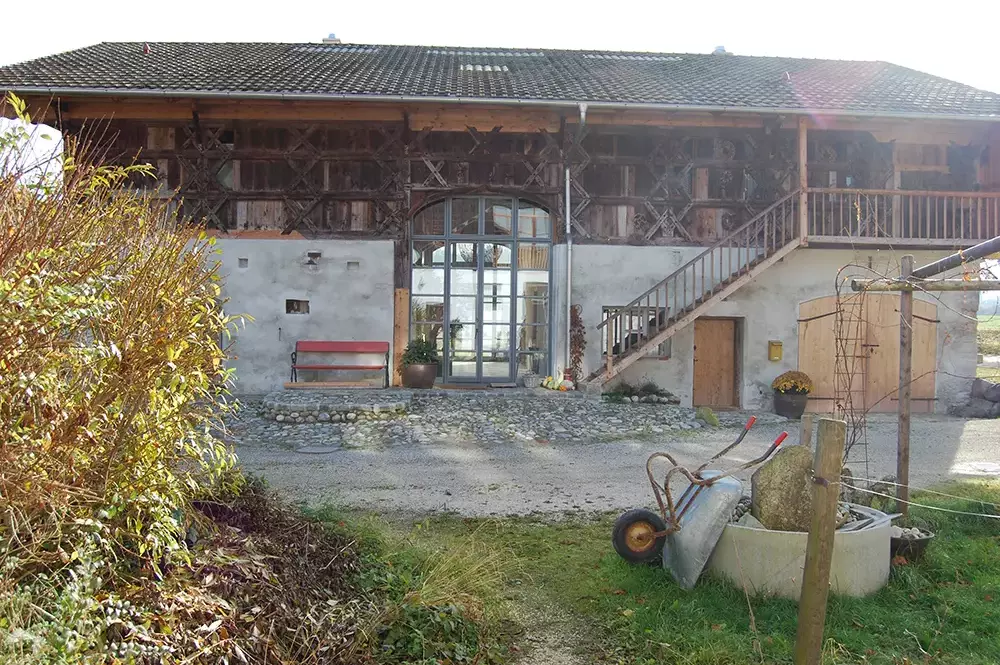 Rustikales Bauernhaus mit Holzfassade, Außentreppe, Steinpflaster und roten Sitzbänken bei Tageslicht.