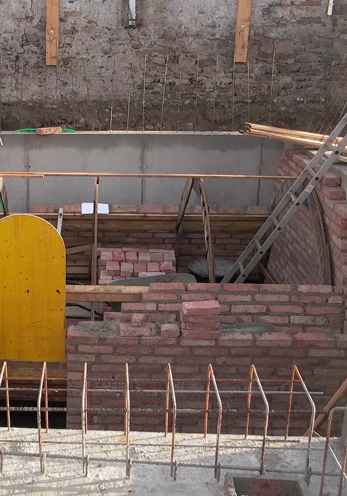 Baustelle mit Mauerwerk aus roten Ziegeln, Holzstützen, Leiter und Bewehrungseisen im Vordergrund.
