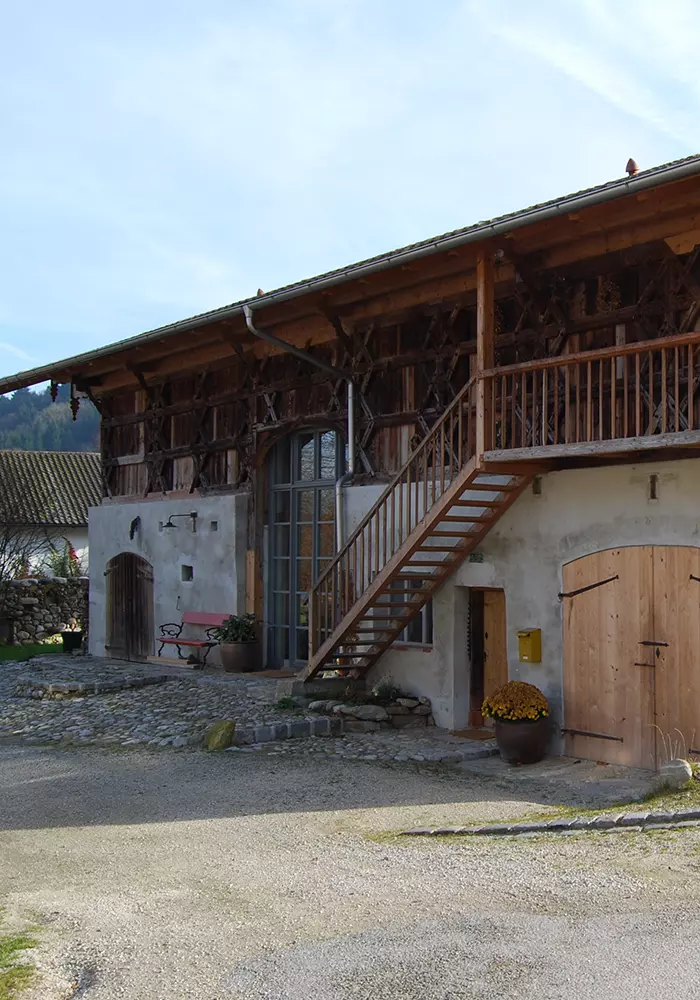 Rustikales Bauernhaus mit Holzfassade, Außentreppe, Steinpflaster und roten Sitzbänken bei Tageslicht.