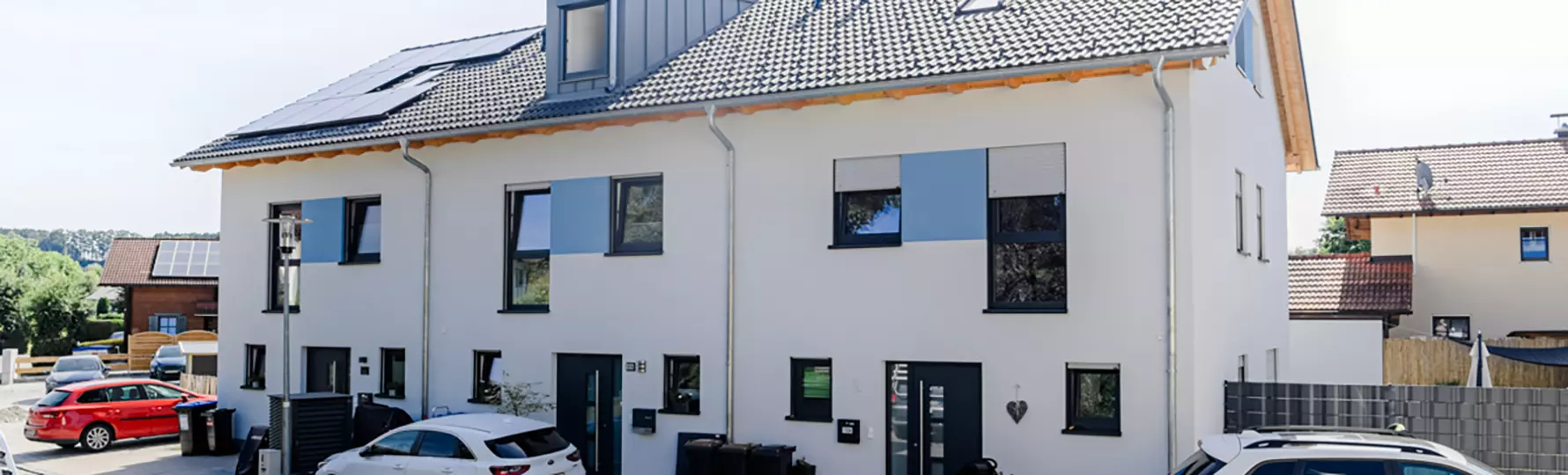 Modernes, frisch errichtetes Doppelhaus mit heller Fassade, dunklen Fenstern, blauen Akzenten und Solarpanels auf dem Dach in einer ruhigen Wohnsiedlung.