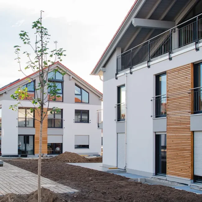 Moderne Wohngebäude in einem Neubaugebiet mit weißer Fassade, großen Fenstern und Holzelementen an der Fassade; der Außenbereich ist noch im Bau mit unbepflanzter Erde und Pflastersteinen.