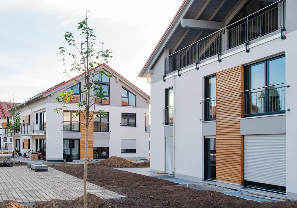 Moderne Wohngebäude in einem Neubaugebiet mit weißer Fassade, großen Fenstern und Holzelementen an der Fassade; der Außenbereich ist noch im Bau mit unbepflanzter Erde und Pflastersteinen.