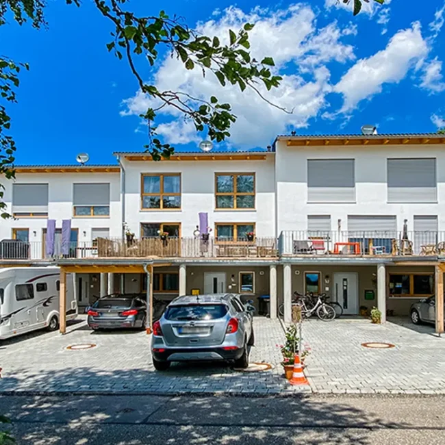 Moderne Reihenhausanlage mit weißer Fassade und Holzdetails, darunter Carports mit parkenden Autos und Wohnmobil, blauer Himmel mit vereinzelten Wolken.