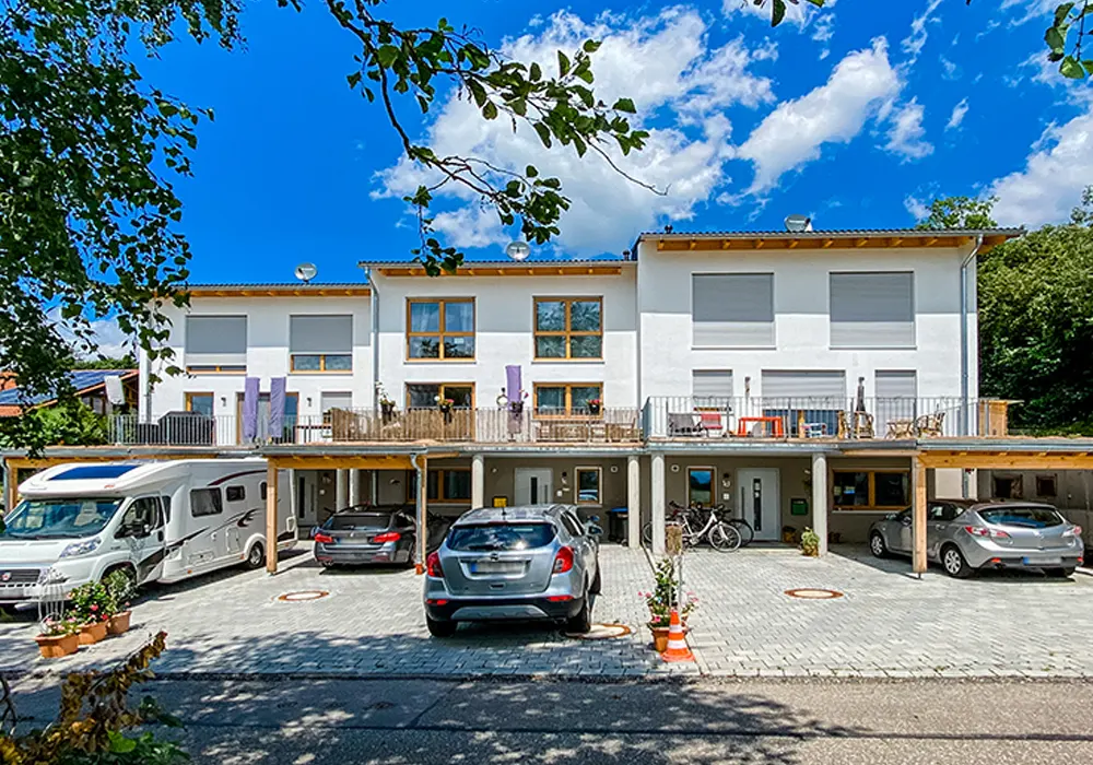 Moderne Reihenhausanlage mit weißer Fassade und Holzdetails, darunter Carports mit parkenden Autos und Wohnmobil, blauer Himmel mit vereinzelten Wolken.