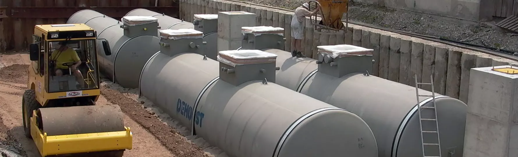 Baustelle mit gelbem Straßenwalzer und mehreren grauen Erdtanks, ein Arbeiter befüllt eine Maschine mit Beton.