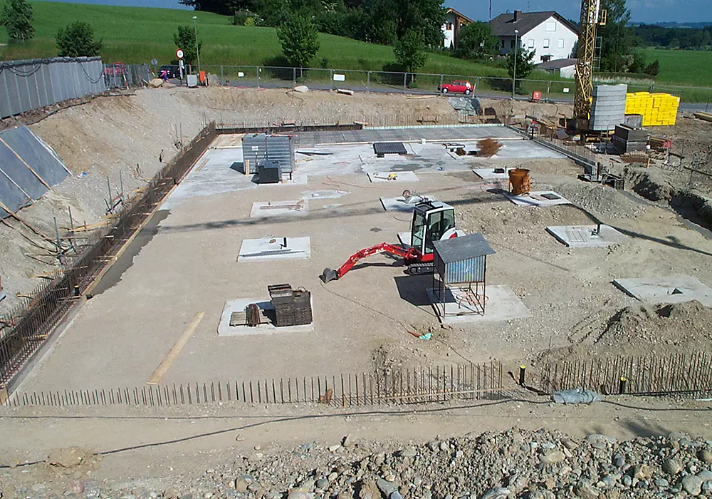 Aushub einer Baugrube mit Fundamentplatte, Bagger und Baumaterialien auf Baustelle im Grünen.
