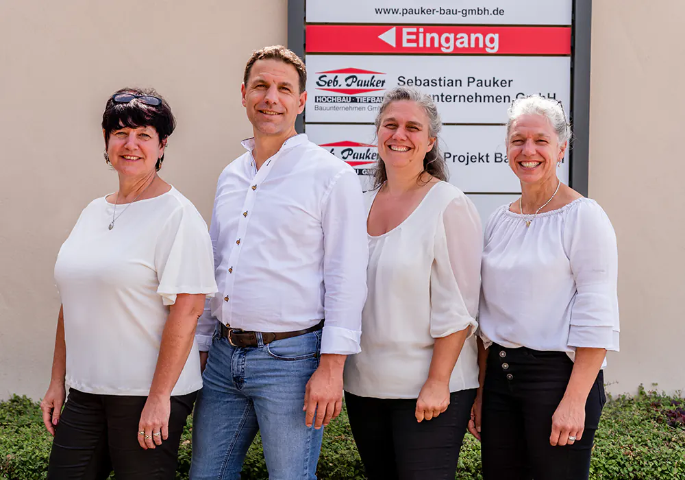 Vier Personen – Maria, Sebastian, Renate, Ingrid – stehen lächelnd vor einem Unternehmensschild mit der Aufschrift ‚Sebastian Pauker Bauunternehmen GmbH‘.