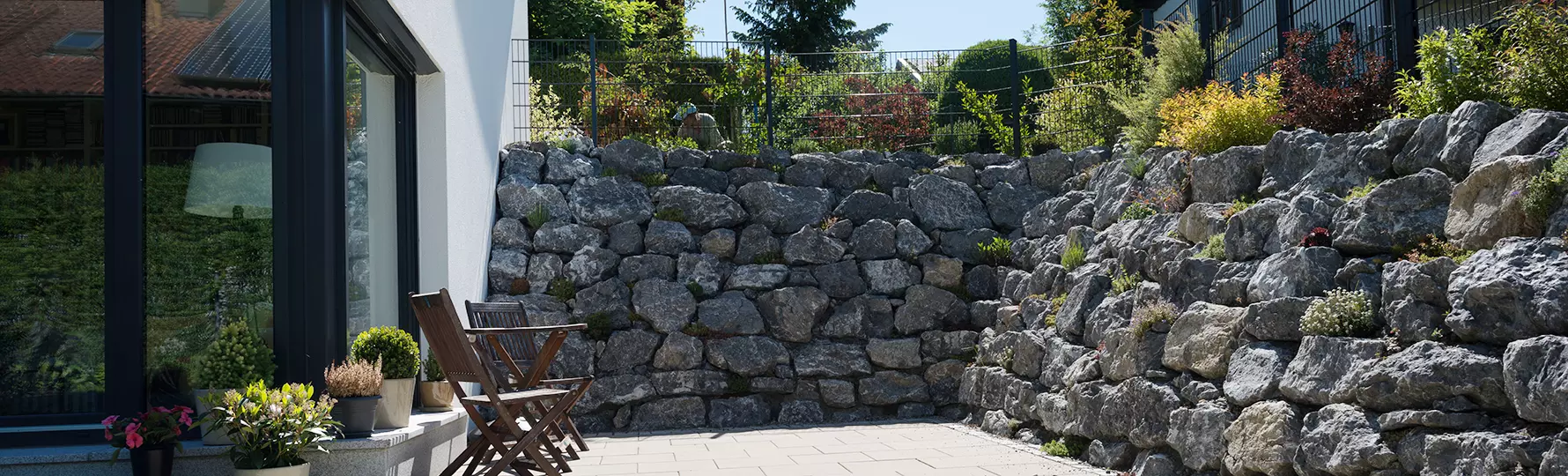 Terrasse mit Holzstühlen vor einem modernen Haus, umgeben von bepflanzten Natursteinmauern als Hangbefestigung.