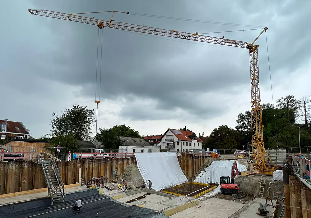 Baustelle mit Baugrube, Kran und Folien an den Wänden; Arbeiter und Bagger bei bewölktem Himmel im Einsatz.