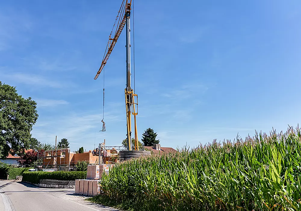 Baukran hebt Bauteil neben Maisfeld auf eine Baustelle mit Rohbau unter blauem Himmel.