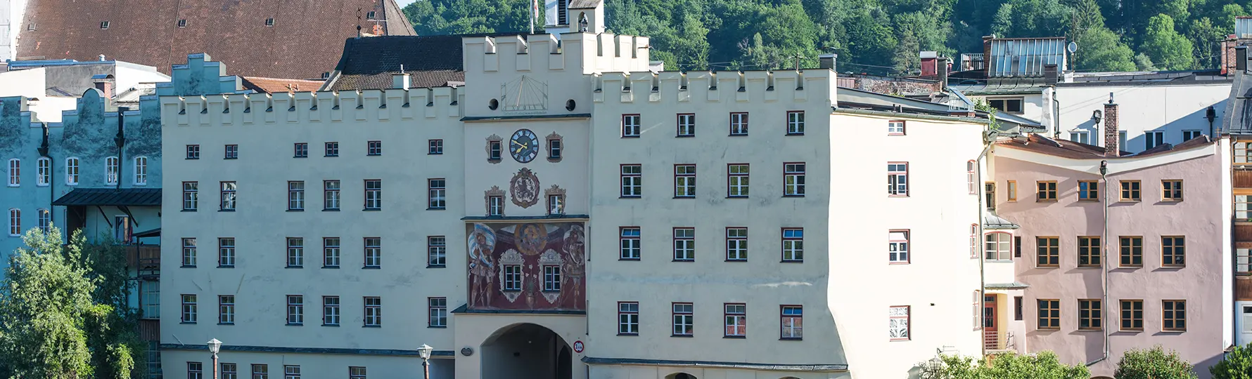 pauker-bau-gmbh-umbau-sanierung-putz-header Historisches Gebäude mit Uhrturm und bemalter Fassade an Flussufer, davor eine überdachte Holzbrücke.