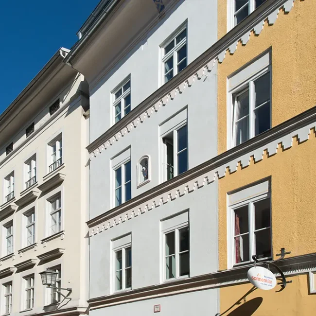 Reihe historischer Stadtgebäude mit weißen und gelben Fassaden, Zierleisten und vielen Fenstern bei blauem Himmel.