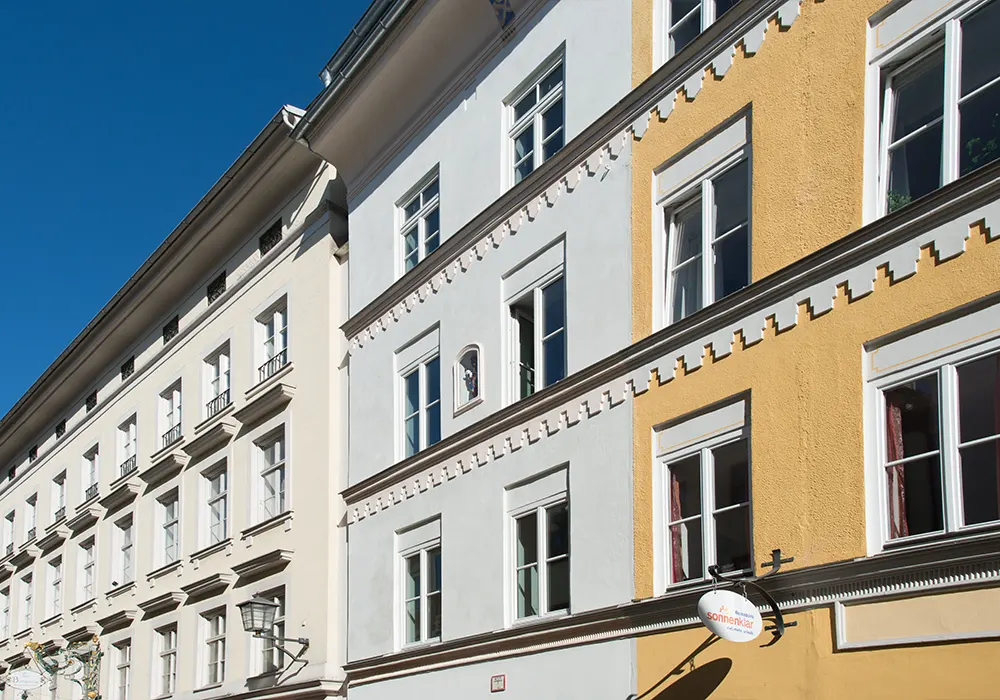 Reihe historischer Stadtgebäude mit weißen und gelben Fassaden, Zierleisten und vielen Fenstern bei blauem Himmel.