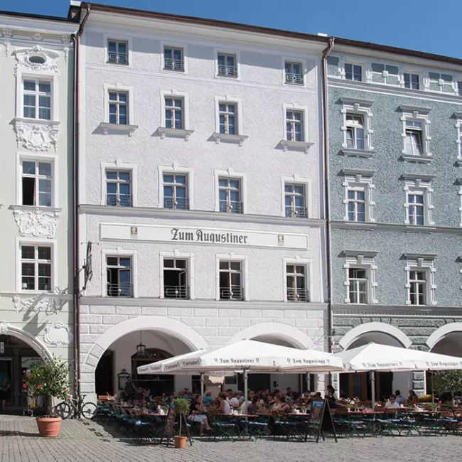 Historisches Gebäudeensemble mit Restaurant „Zum Augustiner“, weißer Fassadenputz, Rundbogenarkaden und Außenbestuhlung auf Kopfsteinpflaster unter Sonnenschirmen.