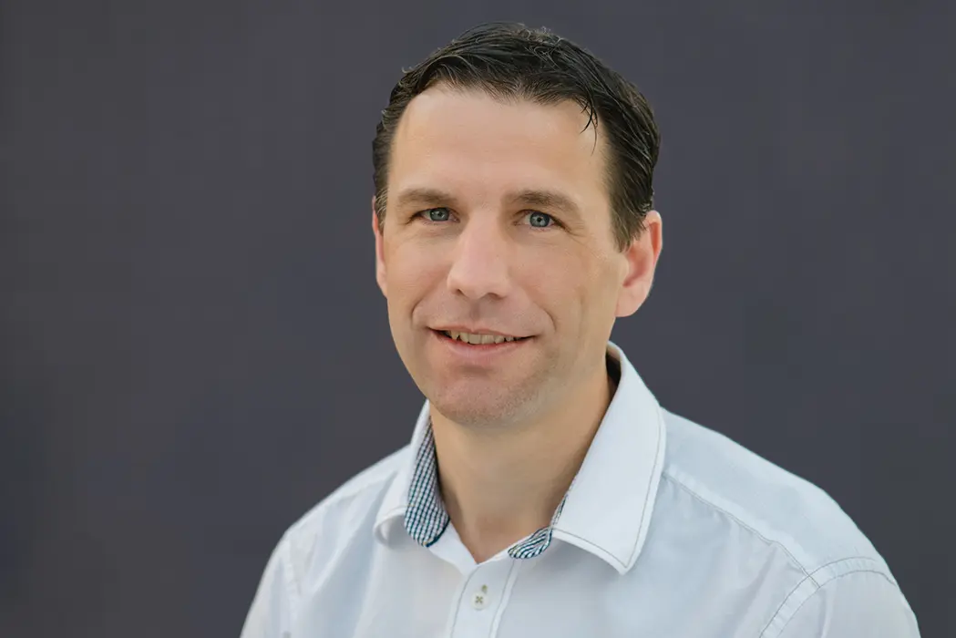 Porträt von Sebastian Pauker mit weißem Hemd vor dunklem Hintergrund, er lächelt leicht in die Kamera.