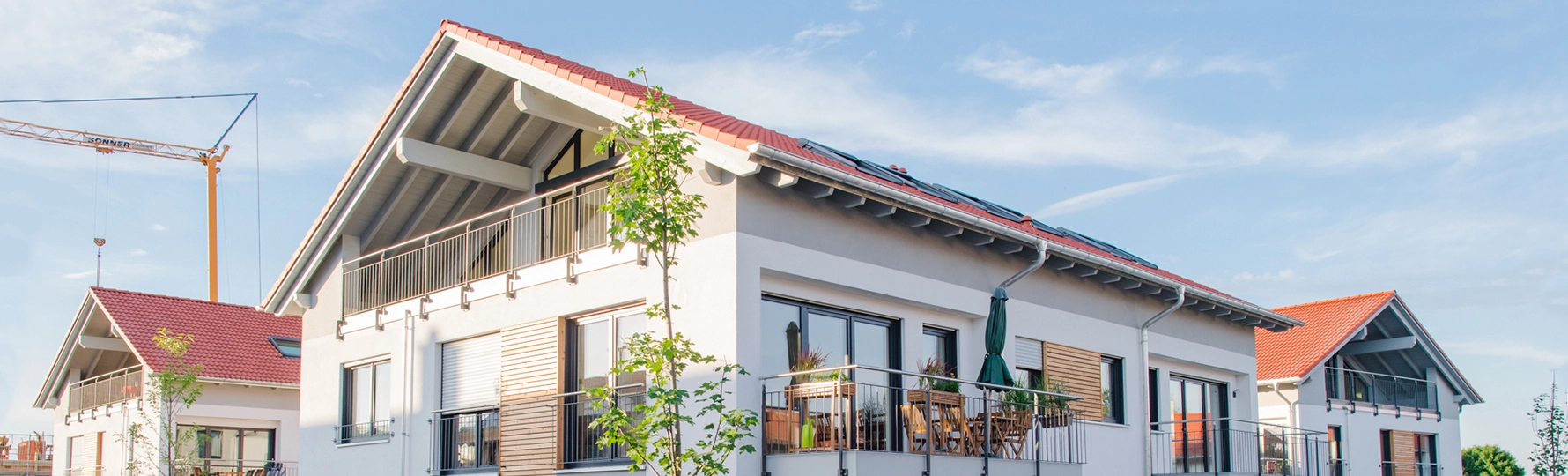 Moderne Mehrfamilienhäuser mit roten Satteldächern, Balkonen und Sonnenschirmen, im Hintergrund ein gelber Baukran.