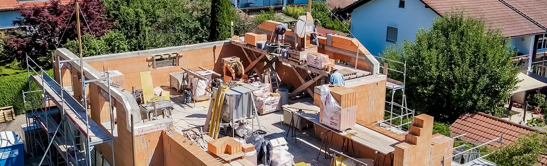 Rohbau eines Einfamilienhauses mit sichtbaren Ziegelwänden und Baustellengeräten, eingerüstet und inmitten einer Wohnsiedlung mit Satteldächern und viel Grün.