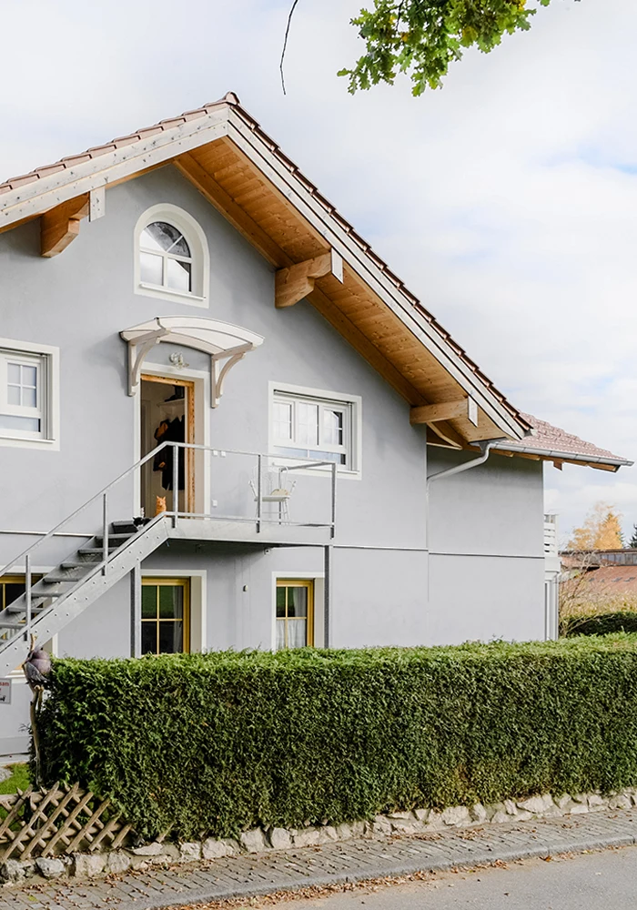 Eingangsbereich eines erweiterten Wohnhauses mit Außentreppe, Balkon und Hecke entlang der Straße