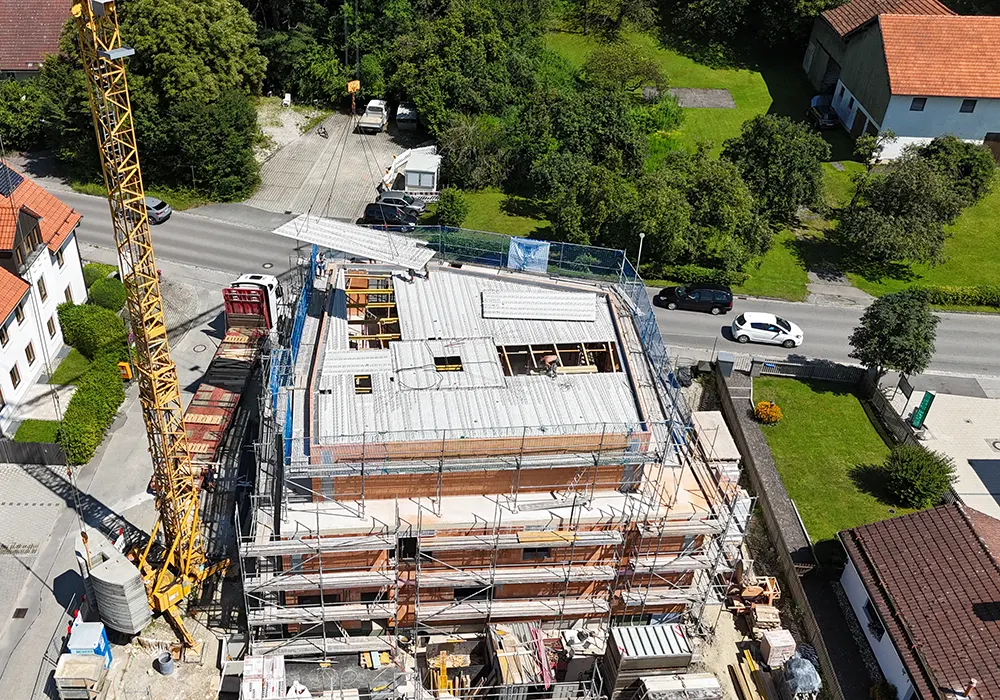 Luftaufnahme eines Rohbaus mit Gerüst und Baustellenkran, Betondecken sind bereits montiert, umgeben von Straße und Häusern.