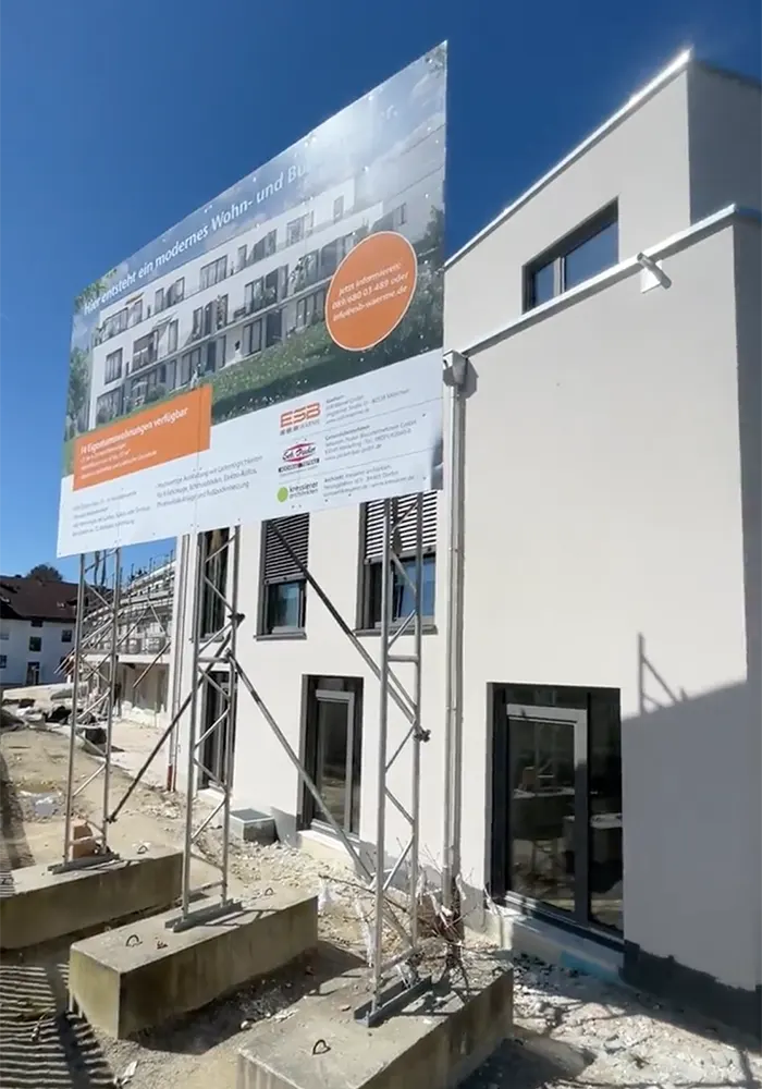 Bauschild vor Neubau mit weißer Fassade und großen Fenstern, Text wirbt für moderne Wohn- und Büroeinheiten.