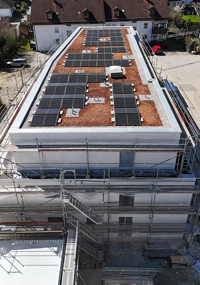 Flachdach mit Solarpanels auf Kiesfläche, eingerüstetes Gebäude im Bau, Wohnhäuser im Hintergrund.
