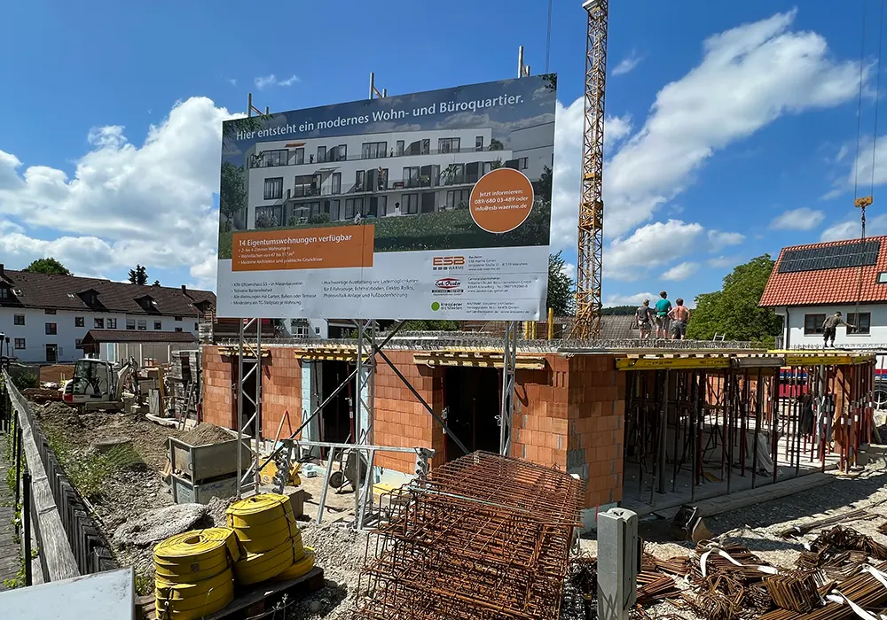 Bauschild vor Rohbau aus Ziegeln mit Baustahl und gelagertem Material, mehrere Personen arbeiten auf der Decke.