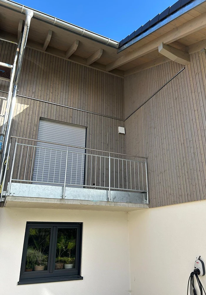 Hausfassade mit Holzverkleidung und Balkon mit Metallgeländer, darunter Fenster mit Pflanzen, rechts Wallbox.