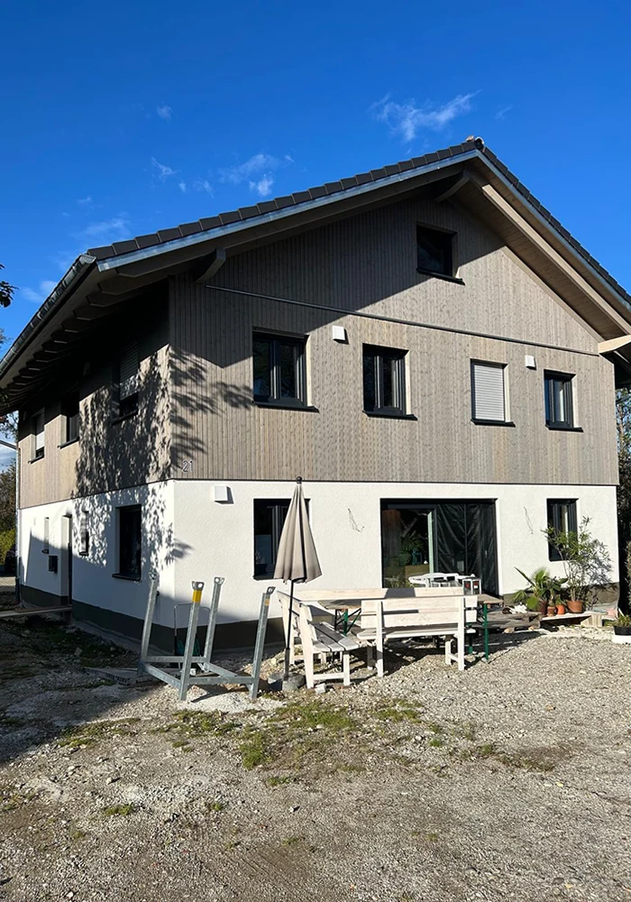 Zweistöckiges Haus mit Holz- und Putzfassade, davor Sitzgruppe mit Sonnenschirm, Schotterboden und Pflanzen.