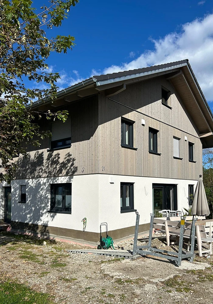 Zweistöckiges Haus mit Holz- und Putzfassade, davor Sitzgruppe mit Sonnenschirm, Schotterboden und Pflanzen.