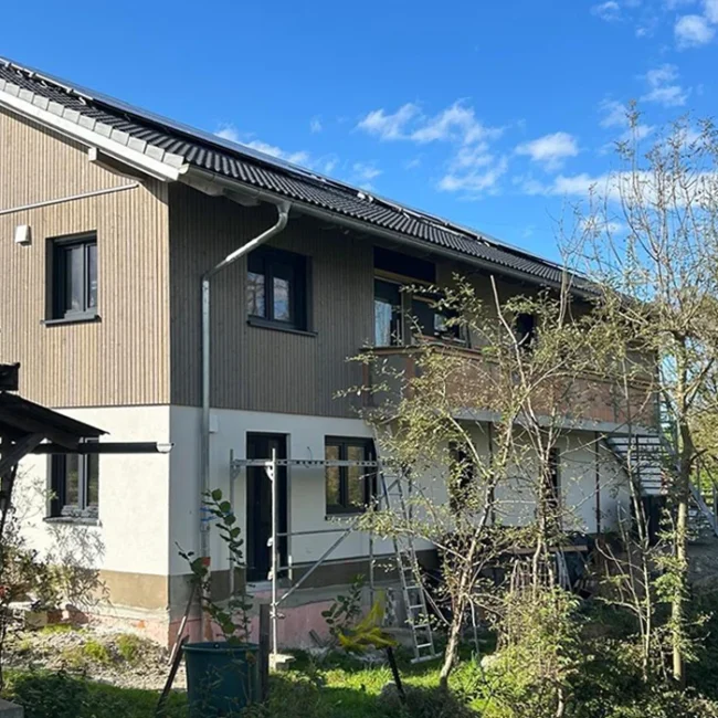 Zweistöckiges Haus mit Holzfassade, sichtbaren Solarpanels und Baugerüst, umgeben von Bäumen unter blauem Himmel.