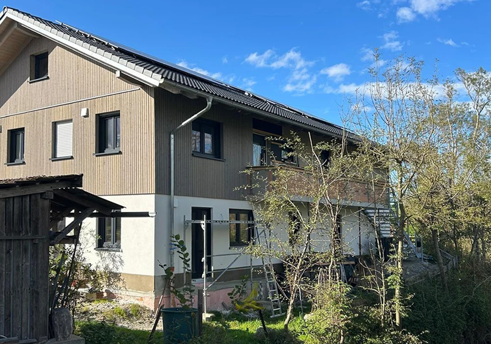 Zweistöckiges Haus mit Holzfassade, sichtbaren Solarpanels und Baugerüst, umgeben von Bäumen unter blauem Himmel.